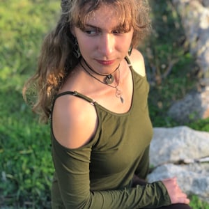 Può includere: Un top verde oliva a maniche lunghe con spalle scoperte. Il top è abbinato a pantaloni marrone scuro. La persona indossa una collana girocollo nera con un ciondolo e orecchini abbinati. Lo sfondo è sfocato con erba verde e rocce.