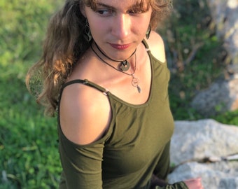 May include: An olive green long-sleeve top with cold shoulder cutouts. The top is paired with dark brown pants. The person is wearing a black choker necklace with a pendant and matching earrings. The background is blurred with green grass and rocks.