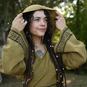 Puede incluir: Top con capucha verde oliva con intrincados patrones geométricos negros en las mangas y el dobladillo. La parte superior presenta un diseño envolvente y está hecha de una tela texturizada. La capucha está levantada.