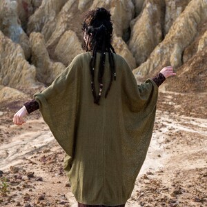 May include: A woman wearing a long, olive green, knitted cardigan with a loose, flowing silhouette. The cardigan has a relaxed fit and long sleeves. The woman has long, dark hair with beads woven into it.