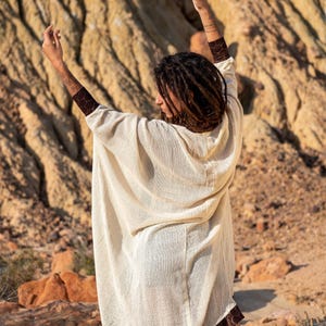 May include: Cream-colored, hooded poncho with a brown geometric patterned trim. The poncho is made of a lightweight, textured fabric. The model is wearing the poncho outdoors in a desert-like environment.