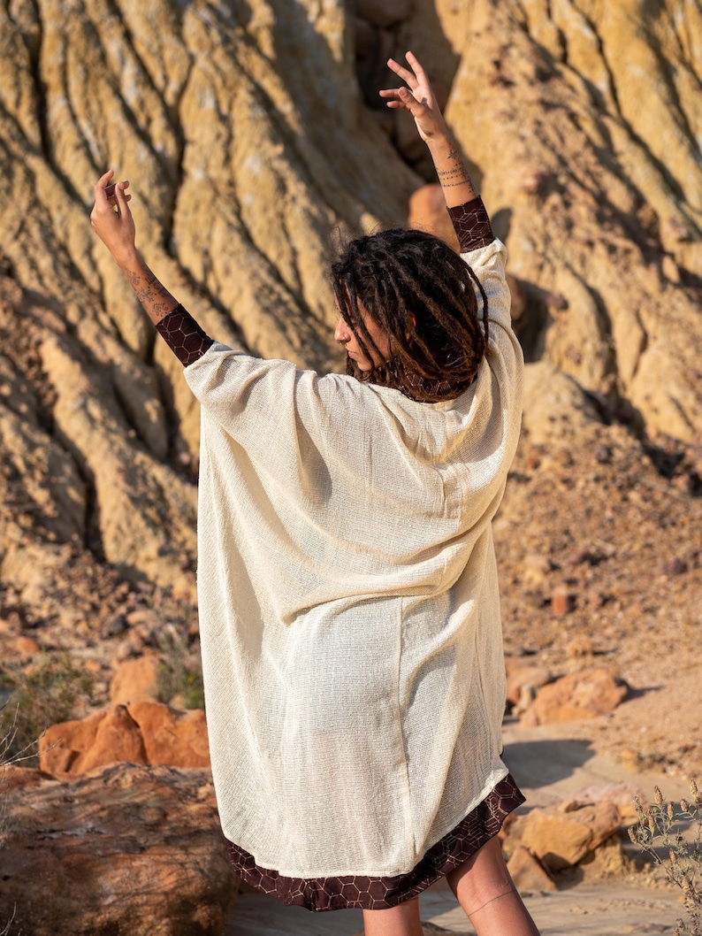 May include: Cream-colored, hooded poncho with a brown geometric patterned trim. The poncho is made of a lightweight, textured fabric. The model is wearing the poncho outdoors in a desert-like environment.