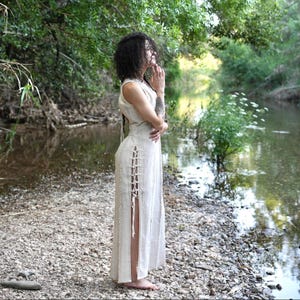 Pode incluir: Um vestido sem mangas creme com fendas laterais e detalhes de cadarço. O vestido é usado ao ar livre perto de um rio, com a pessoa em pé em uma margem rochosa. O vestido tem um decote alto e um design longo até o chão.