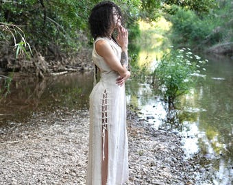 Peut inclure: Une robe sans manches de couleur crème avec des fentes latérales et des détails de laçage. La robe est portée à l'extérieur près d'une rivière, la personne se tenant sur une rive rocheuse. La robe a un col montant et une coupe longue.