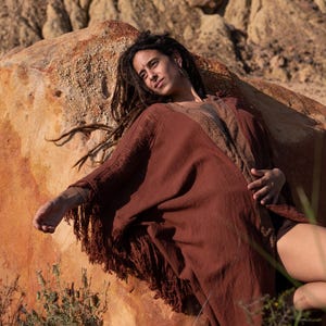 May include: A rust-colored, fringed shawl with a brown patterned trim. The shawl is draped over a person resting on a large, reddish-brown rock. The person has long dreadlocks.