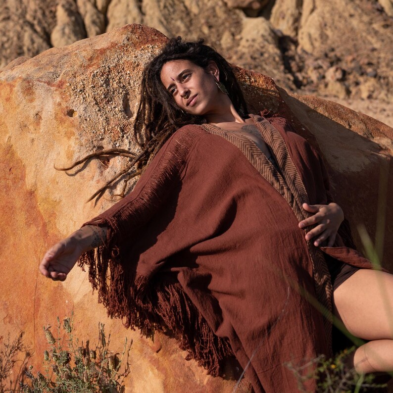 May include: A rust-colored, fringed shawl with a brown patterned trim. The shawl is draped over a person resting on a large, reddish-brown rock. The person has long dreadlocks.