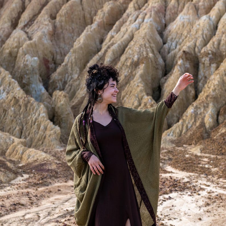 May include: A woman wearing a long, olive green cardigan with a brown dress underneath. She is standing in front of a rocky, textured background.