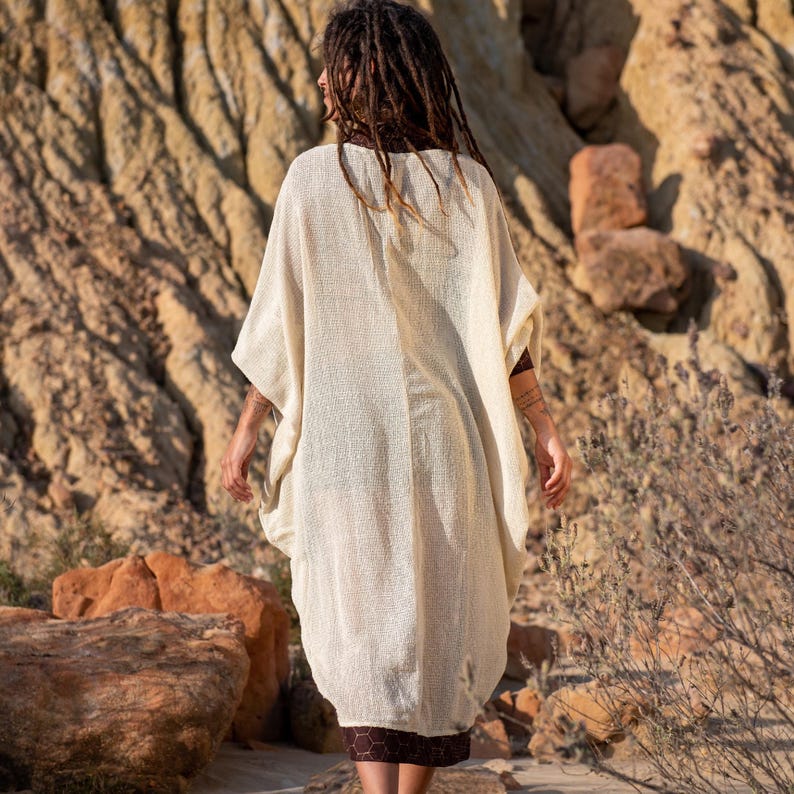 May include: A cream-colored, open-front poncho-style garment with wide sleeves. The poncho has a loose, flowing fit and a textured appearance. The bottom hem of the garment is a dark brown, patterned fabric. The person is standing outdoors.