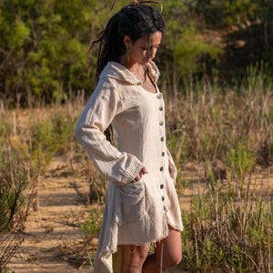 May include: A white linen dress with a long sleeve, a hood, and a button-up front. The dress has a pocket on the left side and a frayed hem.