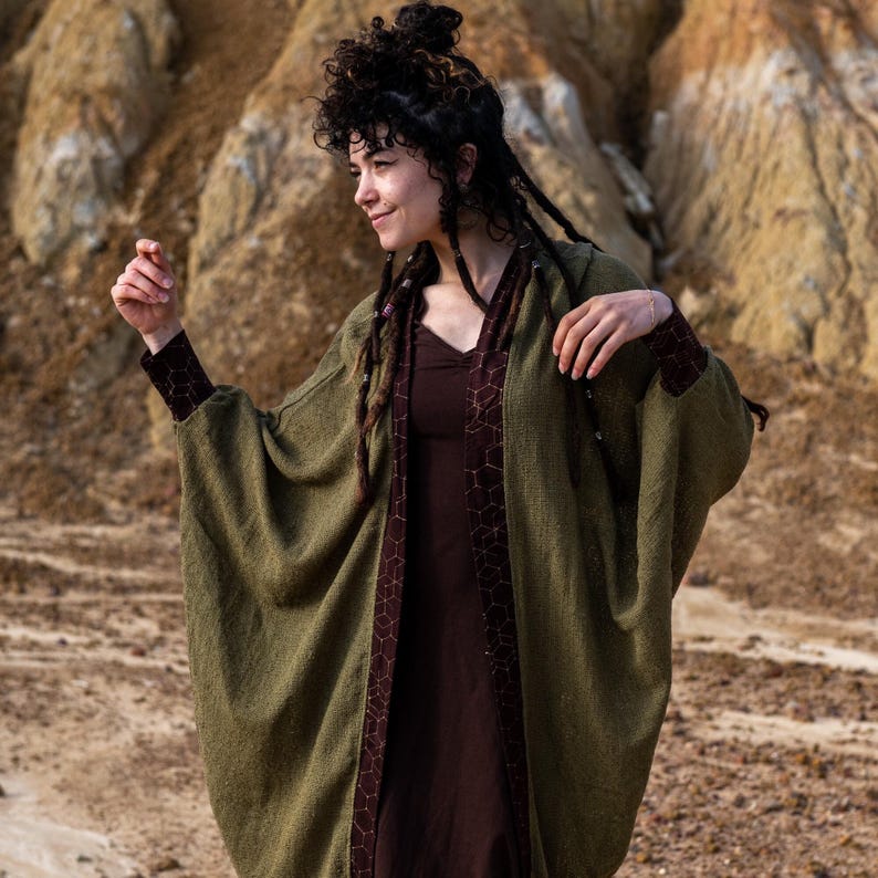 May include: A woman wearing a long, olive green cardigan with a brown geometric pattern on the inside. The cardigan has wide sleeves and a relaxed fit. She is standing in a desert landscape.