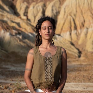 Puede incluir: Una mujer con rastas y un bindi se sienta con los ojos cerrados, meditando en un paisaje desértico. Lleva una blusa verde sin mangas con un patrón geométrico y una falda blanca.