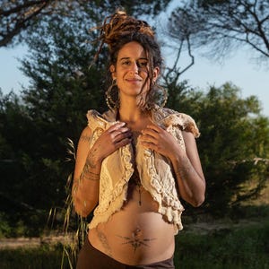 Puede incluir: Una mujer con rastas lleva un chaleco de ganchillo con ribete de encaje. Está sonriendo y mirando a la cámara. Tiene un piercing en el ombligo y un tatuaje en el estómago.