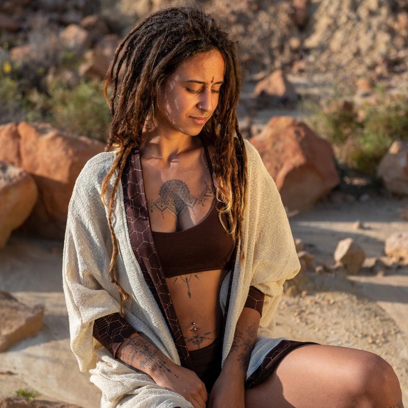 May include: A woman wearing a brown crop top and shorts, with a cream-colored shawl. The shawl has a brown geometric pattern on the edges. The woman has dreadlocks and tattoos on her chest and arms. The background is a rocky, outdoor setting.