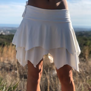 May include: A white, layered skirt with a flowing design. The skirt has a wide waistband and a tiered, asymmetrical hemline. The fabric appears lightweight, suitable for warm weather.