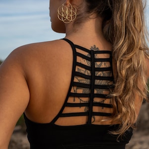 Puede incluir: Sujetador deportivo negro con diseño de espalda con tiras. La persona lleva pendientes de aro dorados. Se ve pelo largo y ondulado y un tatuaje en la espalda. El fondo está borroso, lo que sugiere un entorno al aire libre.
