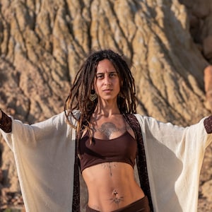 May include: A woman with dreadlocks and tattoos wears a brown crop top and bottoms, with a cream-colored open-front cardigan. The cardigan has brown trim on the sleeves and edges. The background is a tan rock formation.