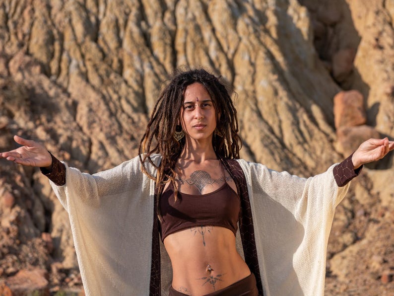 May include: A woman with dreadlocks and tattoos wears a brown crop top and bottoms, with a cream-colored open-front cardigan. The cardigan has brown trim on the sleeves and edges. The background is a tan rock formation.
