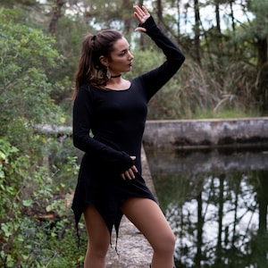 Puede incluir: Una mujer con un vestido negro de manga larga con un dobladillo irregular. El vestido tiene un escote redondo y agujeros para los pulgares en los puños. Está de pie en un entorno boscoso cerca de un cuerpo de agua.