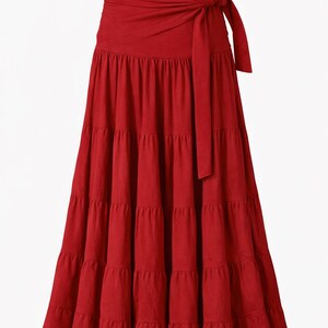 May include: A deep red, tiered maxi skirt with a wrap-around waist and tie closure. The skirt has a flowing, bohemian style. The text "Deep Red" is at the top of the image. The brand name "kāalbelia" is visible on the waistband.
