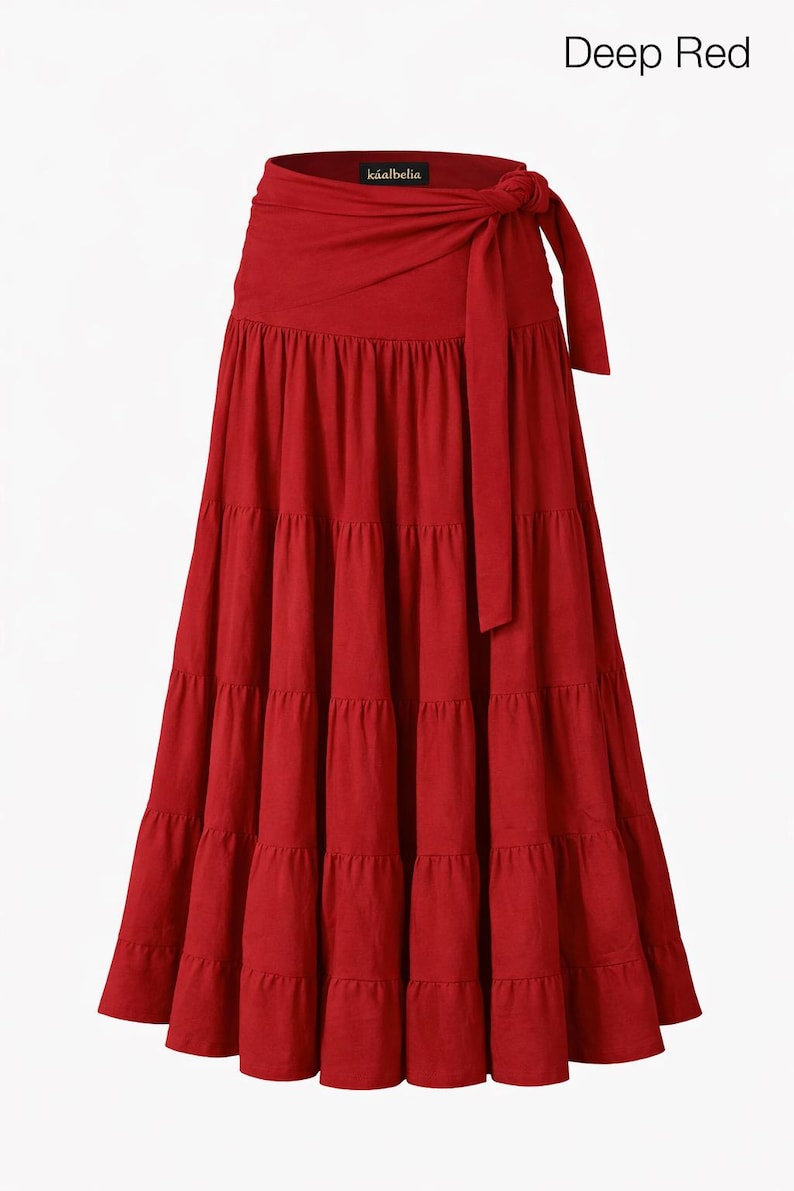 May include: A deep red, tiered maxi skirt with a wrap-around waist and tie closure. The skirt has a flowing, bohemian style. The text "Deep Red" is at the top of the image. The brand name "kāalbelia" is visible on the waistband.