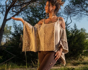 May include: A woman wearing a beige off-the-shoulder top with a geometric pattern and brown pants. The top has a loose, flowing fit and is made of a lightweight fabric. The woman is standing in a field with trees in the background.