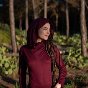 Può includere: Una maglia a maniche lunghe con cappuccio bordeaux. La maglia ha un collo a imbuto e un dettaglio di cucitura a forma di V. La modella indossa la maglia all'aperto con uno sfondo di foresta. La maglia è realizzata in un materiale morbido e confortevole.