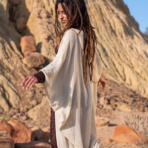 May include: A cream-colored, open-front cardigan with wide sleeves. The cardigan is worn over a brown dress with a geometric pattern. The background features a rocky, desert landscape with warm tones.