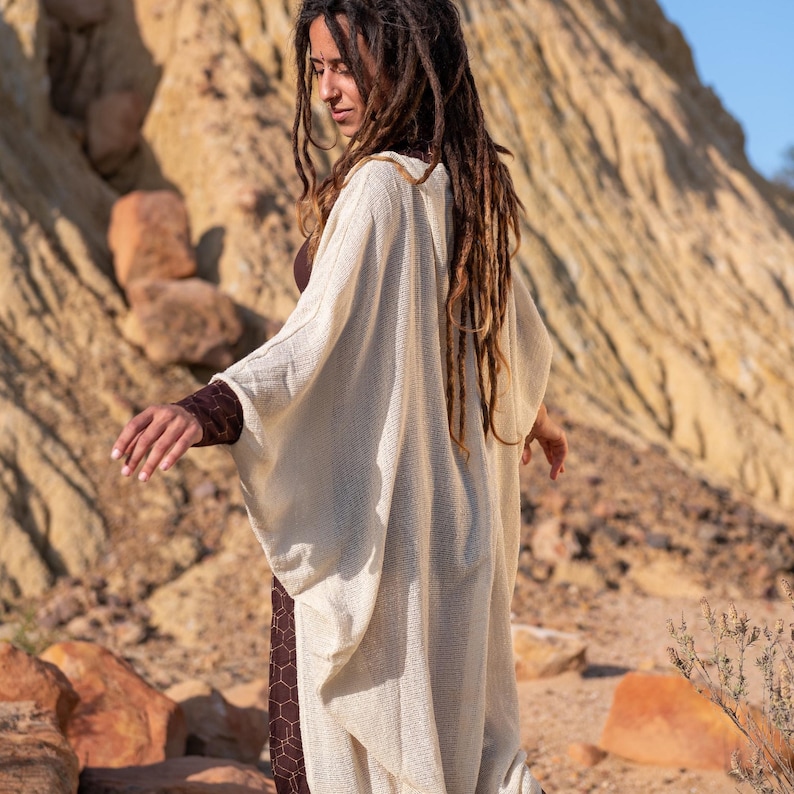 May include: A cream-colored, open-front cardigan with wide sleeves. The cardigan is worn over a brown dress with a geometric pattern. The background features a rocky, desert landscape with warm tones.