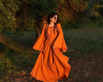 May include: An orange dress with long, flared sleeves and a flowing skirt. The dress features a drawstring neckline and a button-down bodice. The model is standing in a grassy field near a tree, with sunlight filtering through the leaves.
