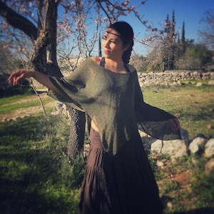 Könnte beinhalten: Ein olivgrüner Strickponcho mit weitem, fließendem Design. Der Poncho wird mit einem langen, braunen Rock kombiniert. Das Bild ist im Freien aufgenommen, mit einem Baum und einer Steinmauer im Hintergrund, unter einem strahlend blauen Himmel.