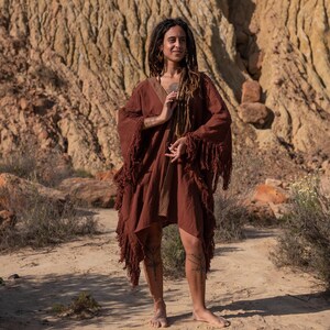May include: A rust-colored, fringed poncho-style garment with a V-neck and decorative trim. The garment is worn outdoors in a desert-like environment. The poncho has long sleeves and a flowing, asymmetrical hemline. The model is barefoot.