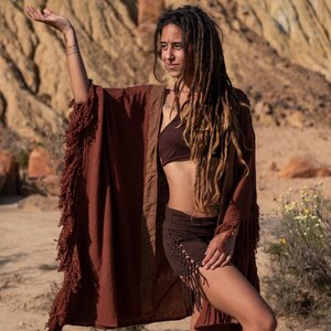 May include: A rust-colored fringed kimono-style robe with a brown chevron pattern on the trim. The robe is paired with a brown bra and shorts. The outfit is suitable for bohemian or festival wear.