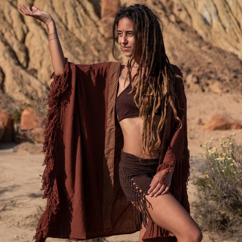 May include: A rust-colored fringed kimono-style robe with a brown chevron pattern on the trim. The robe is paired with a brown bra and shorts. The outfit is suitable for bohemian or festival wear.