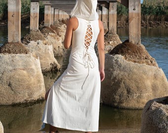 May include: A white hooded dress with a lace-up back detail. The dress has a long, flowing skirt and a fitted bodice. The model is standing on a sandy beach with rocks and water in the background.