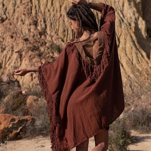 May include: A rust-colored poncho with fringe along the edges and a decorative open back. The poncho has a V-neckline with a patterned trim. The model is wearing the poncho outdoors.