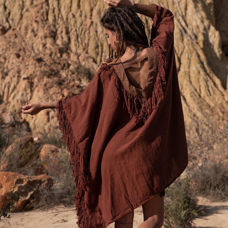May include: A rust-colored poncho with fringe along the edges and a decorative open back. The poncho has a V-neckline with a patterned trim. The model is wearing the poncho outdoors.