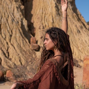 May include: A woman wearing a rust-colored, open-back poncho with fringed edges and a geometric pattern on the shoulders. She has long dreadlocks and is wearing gold-colored earrings. The background features a natural, earthy landscape.
