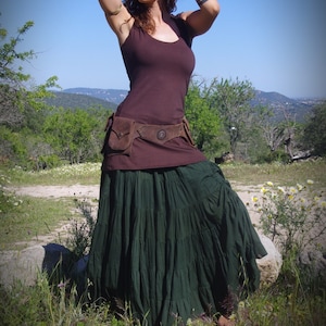 May include: A woman wearing a dark brown tank top, a brown belt with pockets, and a long, dark green tiered skirt. The outfit is worn outdoors with a blue sky and green foliage in the background.