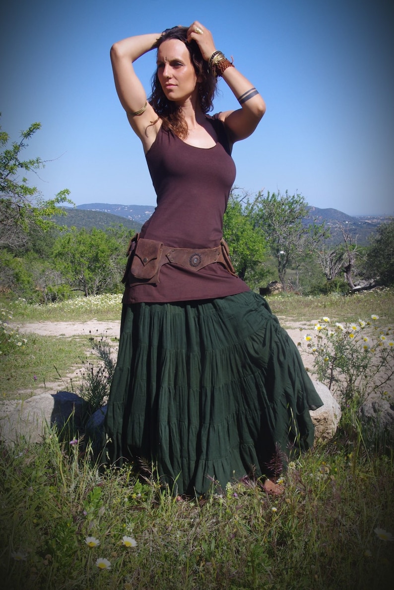 May include: A woman wearing a dark brown tank top, a brown belt with pockets, and a long, dark green tiered skirt. The outfit is worn outdoors with a blue sky and green foliage in the background.