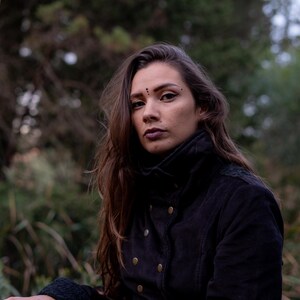 May include: A woman with long brown hair wearing a black jacket with gold buttons. She is standing in a forest setting.