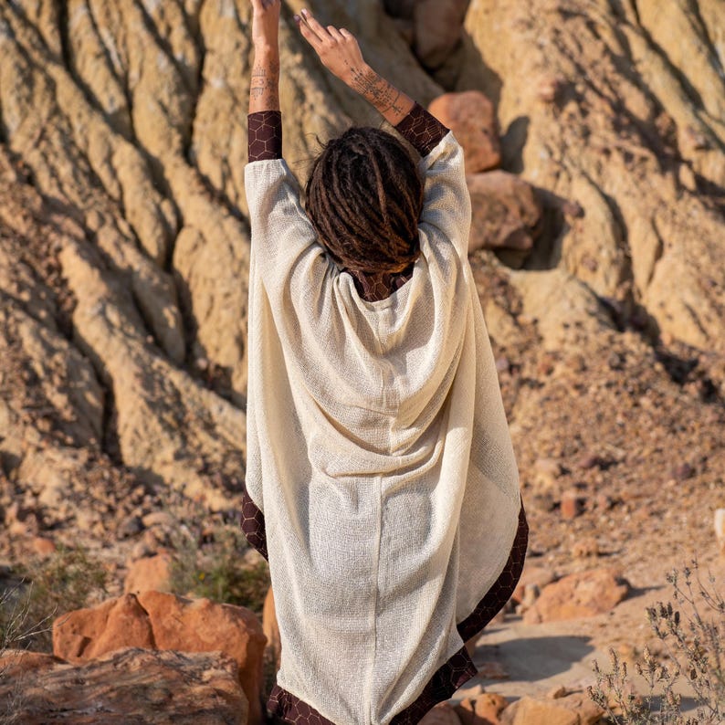 May include: Cream-colored hooded poncho with brown trim. The poncho has a textured appearance and is worn outdoors. The trim features a geometric pattern. The person wearing the poncho has dreadlocks and tattoos on their arms.