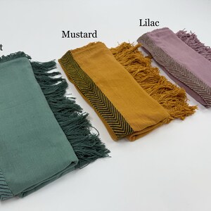 May include: A collection of folded scarves in mint, mustard, and lilac. Each scarf features a fringed edge and a patterned border. The text labels each scarf's color. The scarves are displayed on a white surface.