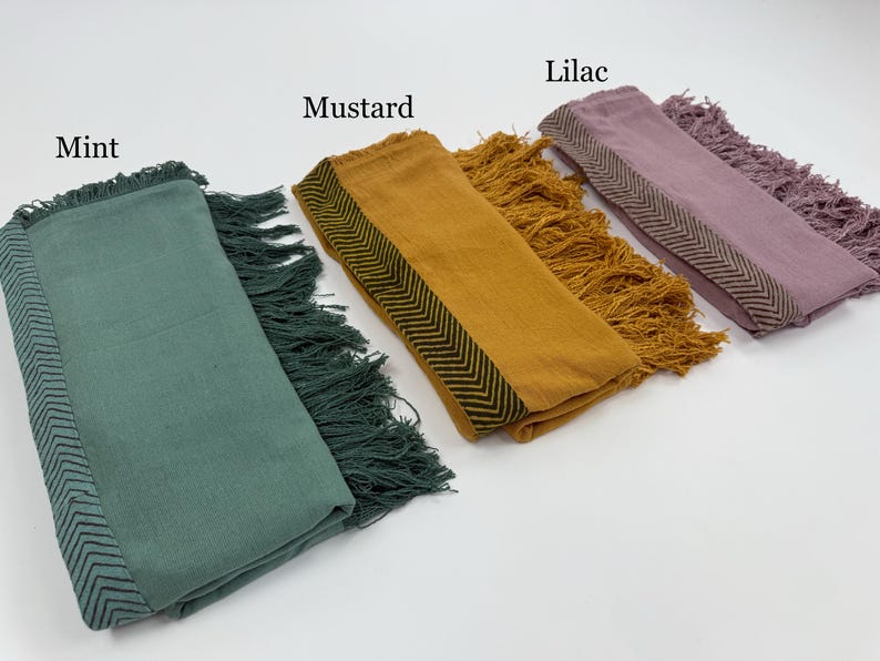 May include: A collection of folded scarves in mint, mustard, and lilac. Each scarf features a fringed edge and a patterned border. The text labels each scarf's color. The scarves are displayed on a white surface.