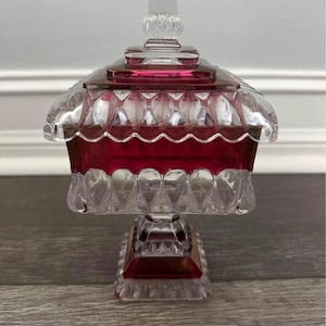 May include: A clear and ruby red glass candy dish with a lid. The dish has a square base and a scalloped edge. The lid has a clear, square finial. The dish is likely vintage.