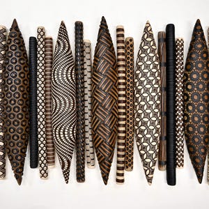 May include: A collection of elongated, spindle-shaped wooden objects and thin cylindrical rods arranged vertically. Each piece features intricate, dark, burned-in geometric patterns, including circles, cross-hatching, waves, and basket-weave designs.
