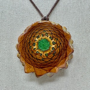 May include: A circular pendant necklace featuring a cross-section of a brown pine cone with a vibrant green resin center. The pendant is suspended from a thin, dark brown cord, showcasing the natural wood grain and textured scales.