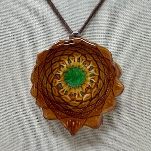 May include: A circular pendant featuring a cross-section of a brown pine cone with a glossy finish and a vibrant green center, suspended from a thin dark brown cord. The natural scale pattern of the pine cone radiates outward from the center.