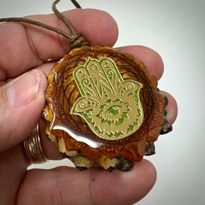 May include: A gold hamsa hand charm pendant with green details, set in a brown resin base with a mandala design.