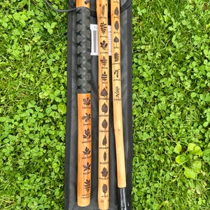 May include: A set of three wooden walking sticks with engraved leaf designs and tree names. The sticks are in a black carrying case on a bed of green grass. The sticks are made of light brown wood with dark brown engravings.