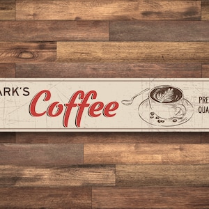 May include: A vintage-style metal sign with a distressed white background and brown text. The sign reads "Mark's Coffee" in red lettering and features a drawing of a cup of coffee with a spoon. The sign also says "Premium Quality!" in brown text.